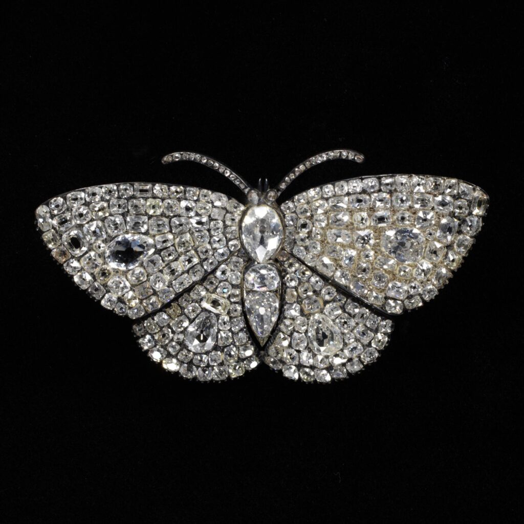 Diamond butterfly shown frontally. Four wings set with massed small diamonds. The body is made up of three larger rose-cut diamonds with two curled proboscis. 