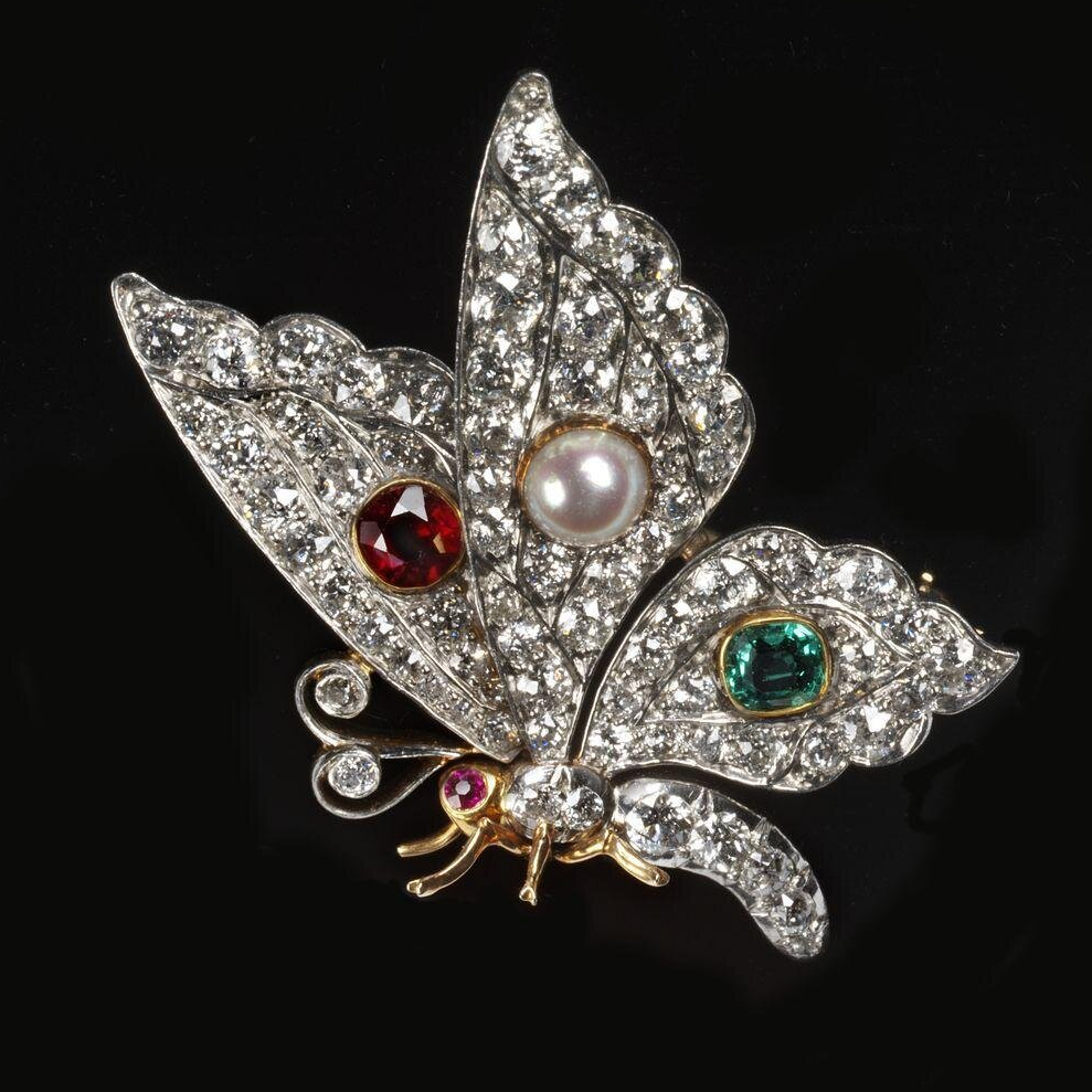 Diamond set butterfly shown facing to the left. Wings are set with, from the left, a ruby, pearl and emerald. The body is made of gold with a ruby eye . The curled proboscis is set with two diamonds. 