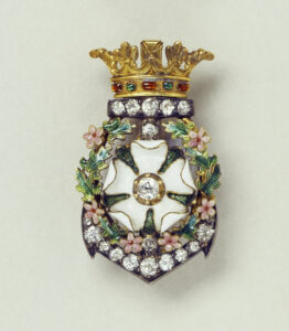 Gold, diamond and enamelled brooch. The upper part is formed of a gold coronet set with alternating rubies and emeralds. Under the coronet, a band of diamonds forms the top part of an anchor. The centre of the jewel is set with a white enamelled rose surrounded by a wreath of green leaves and pink flowers. The base of the jewel is comprised of the bottom part of the anchor, set with diamonds. 