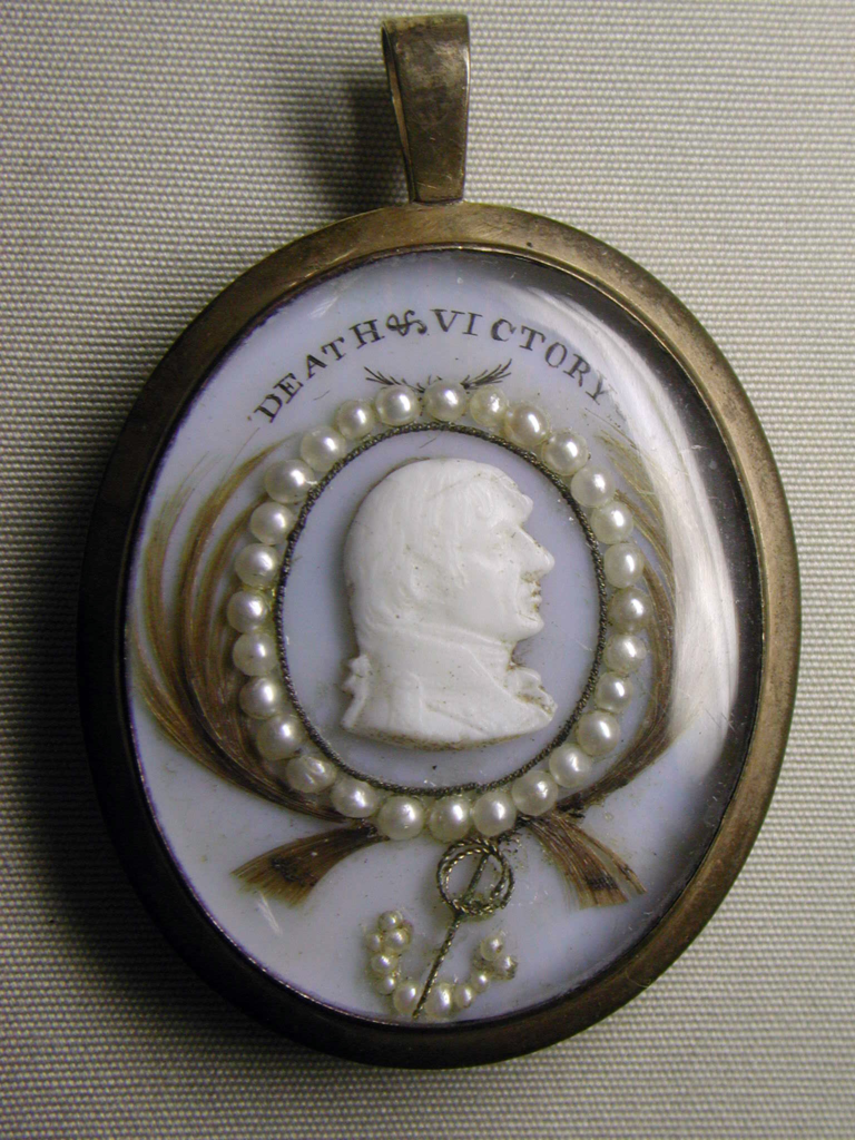 Oval medallion set with glass cameo portrait of Horatio Nelson. Nelson is facing to the right, his hair is combed over his forehead and he wears a high collared shirt. The cameo is set on a light blue background on which the motto Death and Victory is printed. A circle of pearls and locks of hair surrounds the white cameo portrait. A small pearl set anchor hangs from the image. 