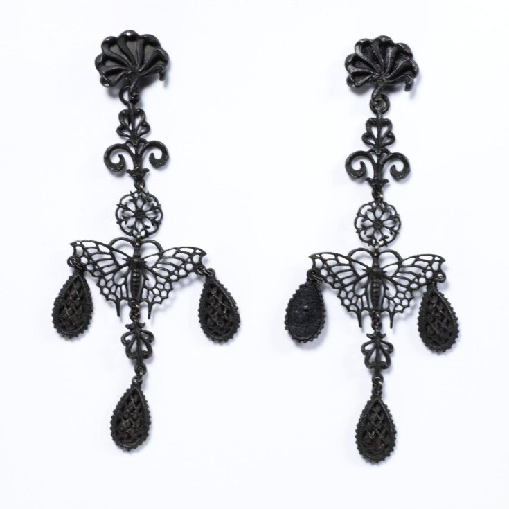 Pair of delicate black cast iron earrings. The top part is formed of a floral rosette from which hang a double scroll and a further openwork rosette. The central part of each earring is made up of a openwork butterfly shown frontally. Three oval pendants hang from the butterfly wings and body. 