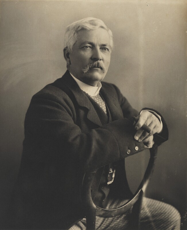 Sepia photograph of explorer Henry Morton Stanley in 1890. Stanley is wearing a dark coat, spotted cravat and light checked trousers. He is seated backwards on a chair with his arms resting on the open back. He has short white hair, a drooping moustache and looks directly at the viewer. The photograph was taken by his sister in law Eveleen Myers.