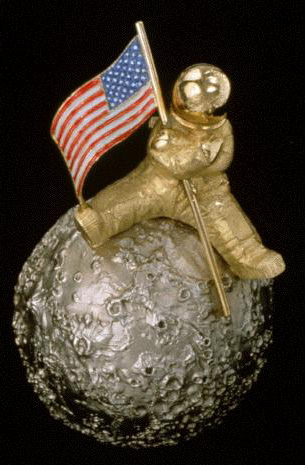 It shows an astronaut in yellow gold holding the red, blue and white American flag decorated with enamel stars, sitting ona white gold moon. The lower part of the flag points towards the Mare Tranquillitatis where Neil Armstrong and Buzz Aldridge first stepped onto the moon. 