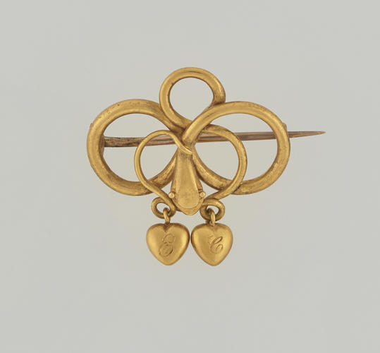 A gold brooch in the form of a snake twisted into three loops, with two engraved hearts suspended. The brooch has a long horizontal pin at the back. 