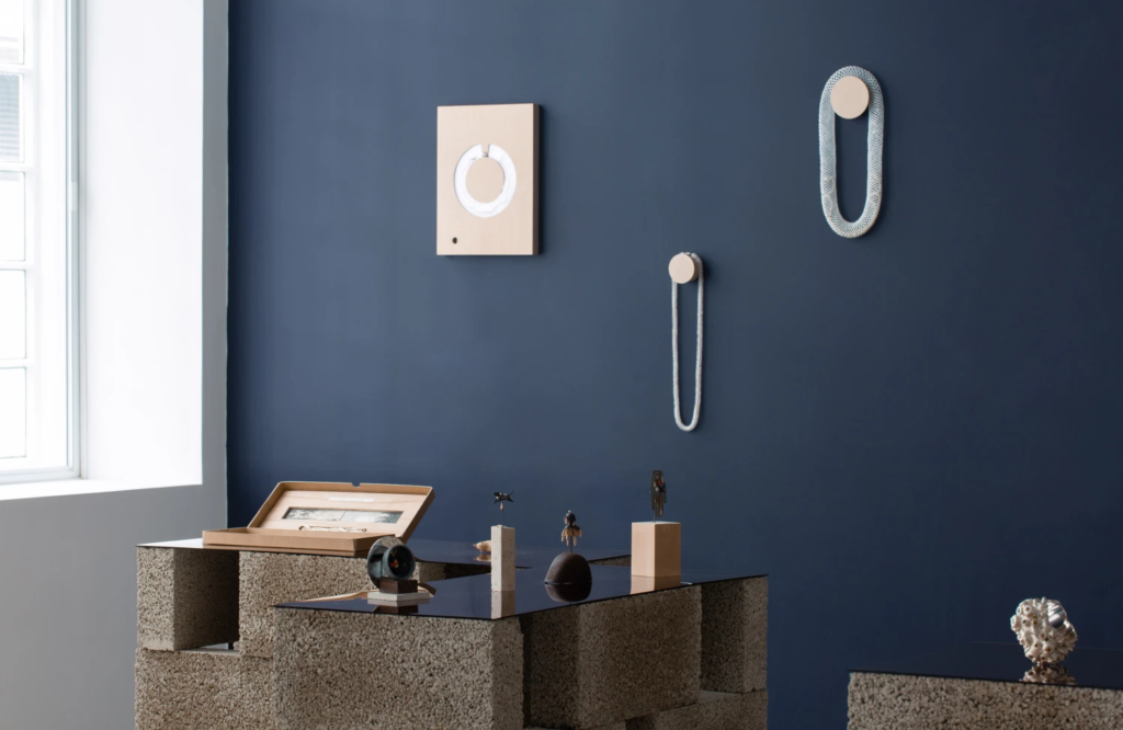 Photograph of the exhibition interior of Memories are made of this, showing necklaces by Caroline Broadhead hanging on a dark blue wall. The breeze block stands with mirrored tops in the foreground show jewels by Zoe Arnold and the silver and citrine bracelet by Silvia Weidenbach. 