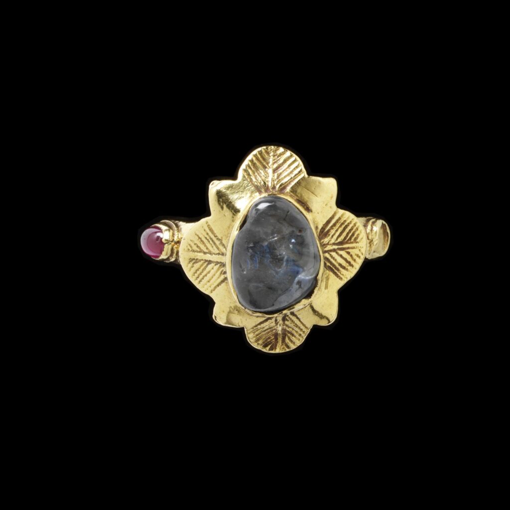 Gold ring with flower shaped bezel set with a round cabochon sapphire in the centre and pink sapphires on each side. Part of Edmund Waterton's ring collection. 