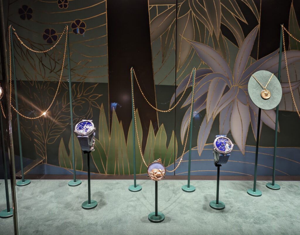 Display of watches and necklaces from the 'Poetry of Time' exhibition by Van Cleef and Arpels, London. Showin two watches and two pendants on green metal mounts on a green case base and a floral background. 