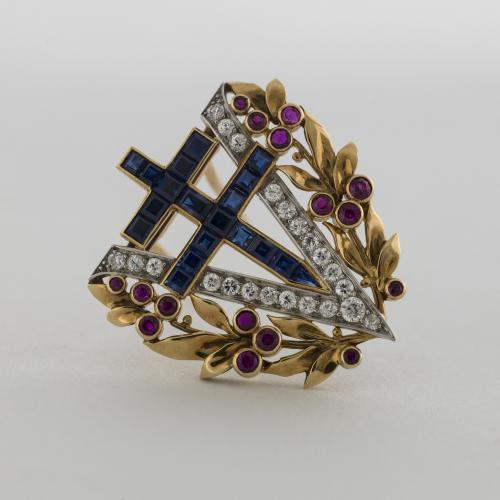 Gold, platinum, diamond, ruby and sapphire brooch in the shape of a capital letter V with a border of gold leaves and red berries. The centre of the V is filled with a sapphire double armed cross. 'Broche de la Victoire' by Van Cleef and Arpels, 1944