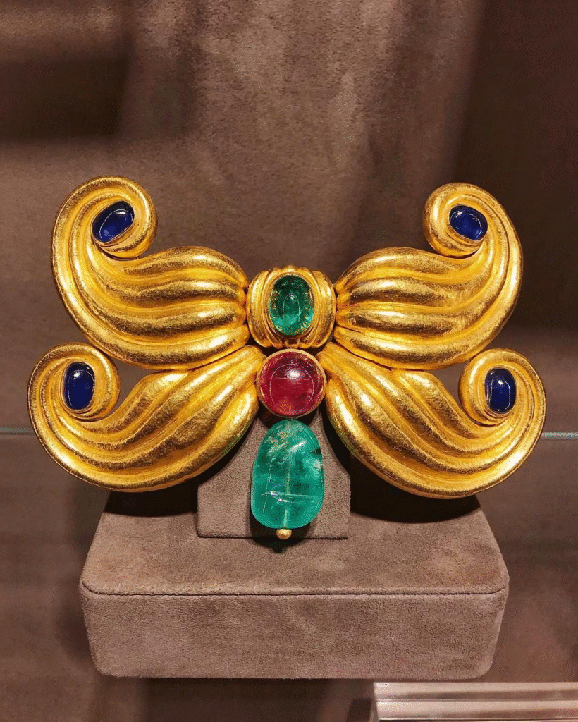 Brooch by Suzanne Belperron shaped like four curls of ridged gold around a cabochon emerald and ruby. The curls of gold are placed one above the other like a double moustache. A pendant cabochon ruby depends from the bottom. 